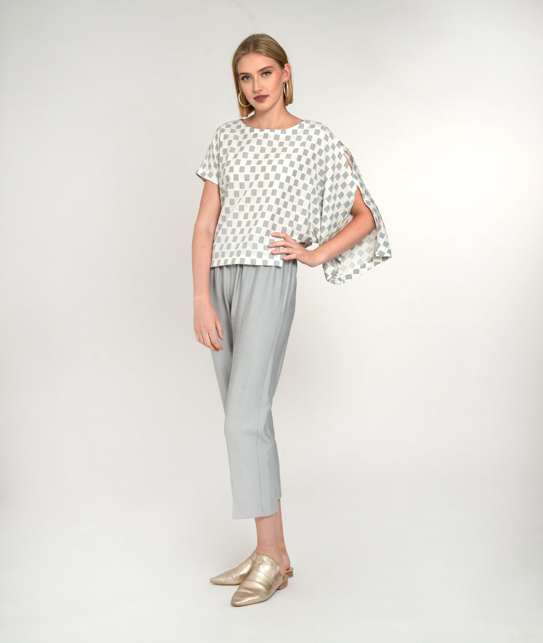 model in a slim cropped grey pant with a grey and white checker board print top, with an asymmetrical body and one long open draped sleeve