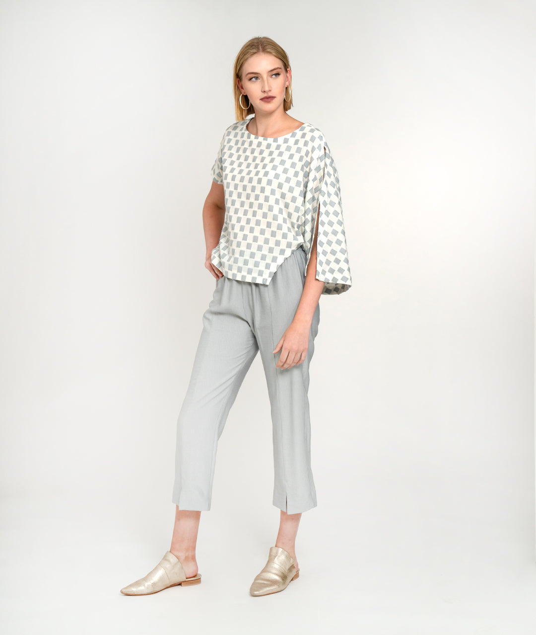 model in a slim cropped grey pant with a grey and white checker board print top, with an asymmetrical body and one long open draped sleeve