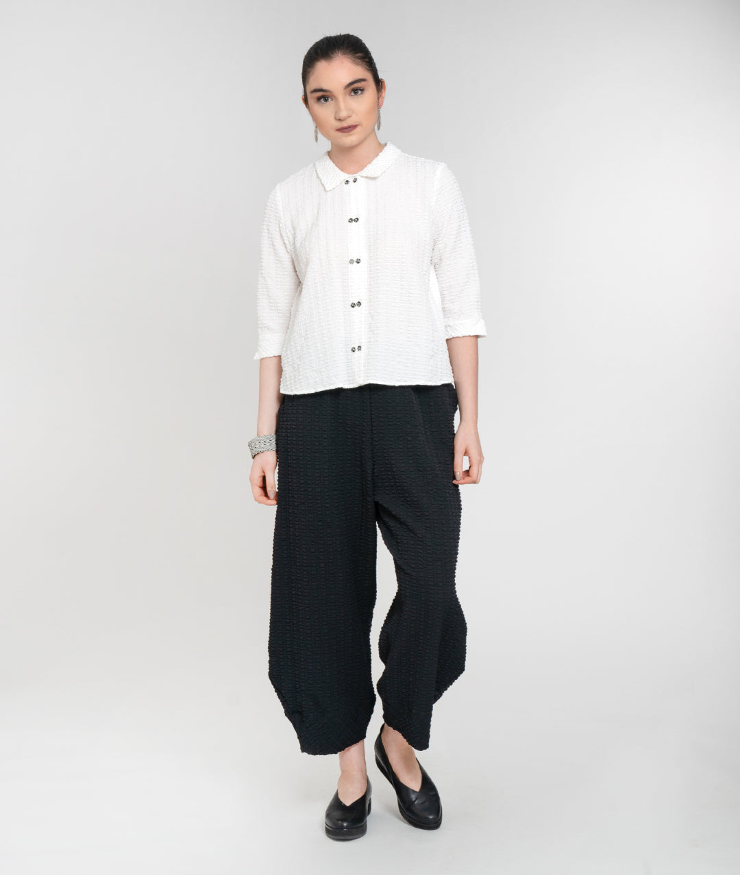  model in a textured white button down blouse. top has 3/4 sleeves and a double button feature up the placket, worn with black pants with a wide leg with a tapered ankle