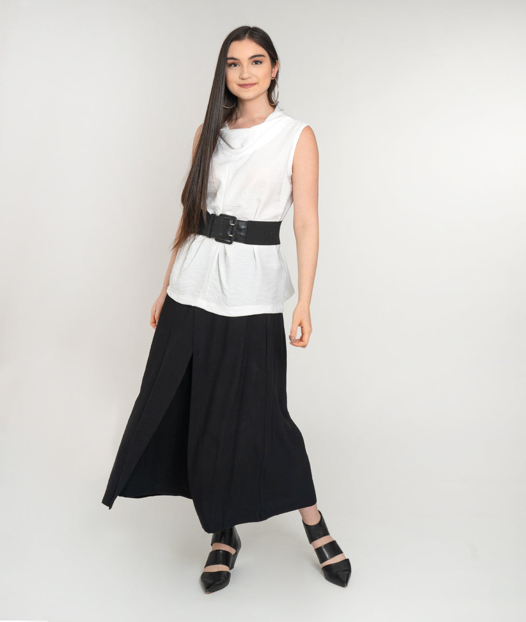 model in a sleeveless white tank with a cowl neck and black belt, worn over a wide leg black pant with an overlapping overlay on either front