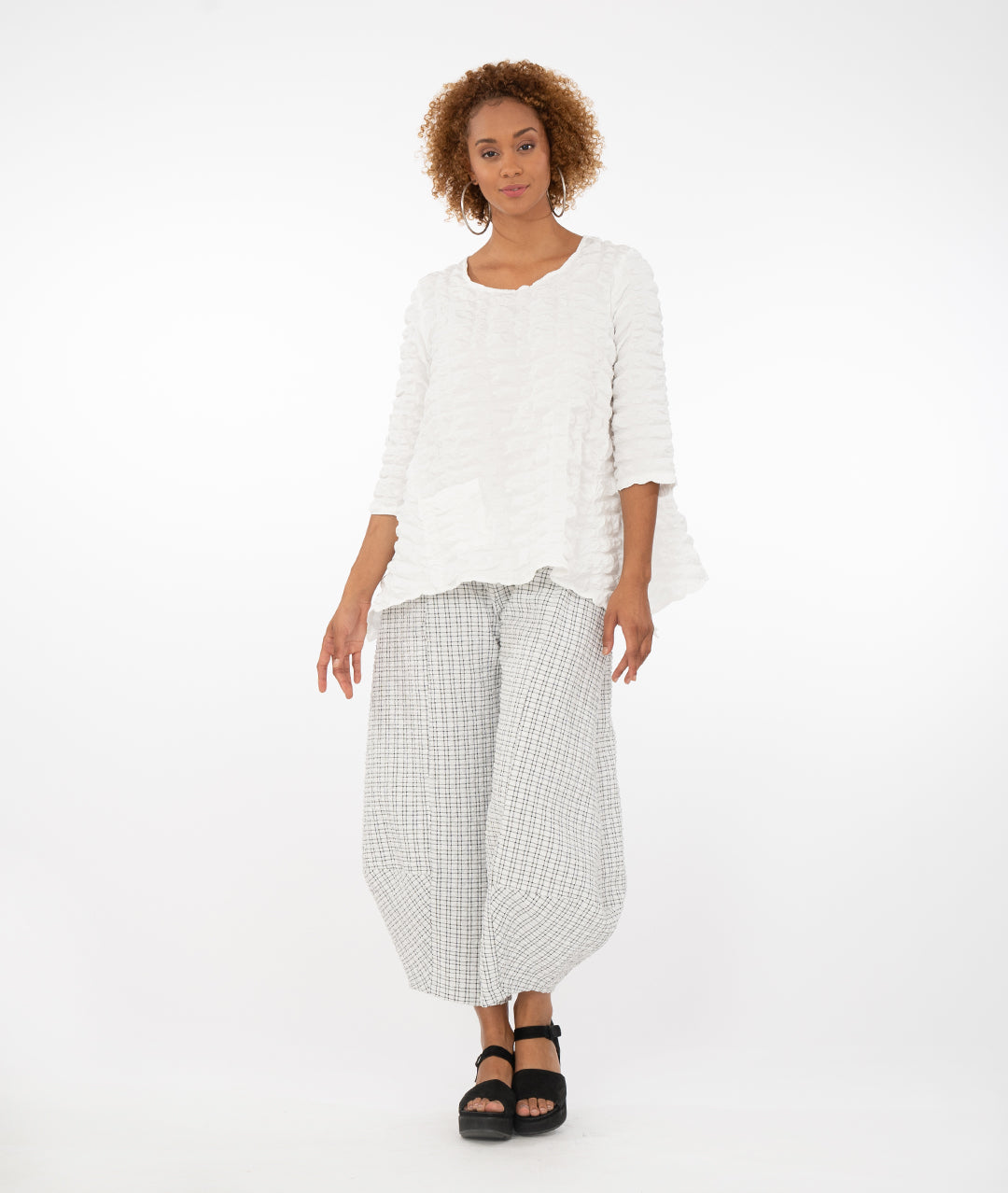 model in a white and black plaid wide leg pant with a tapered ankle, worn with a white pull over top with 3/4 sleeves, dipped hem on either side, and a single square hip pocket