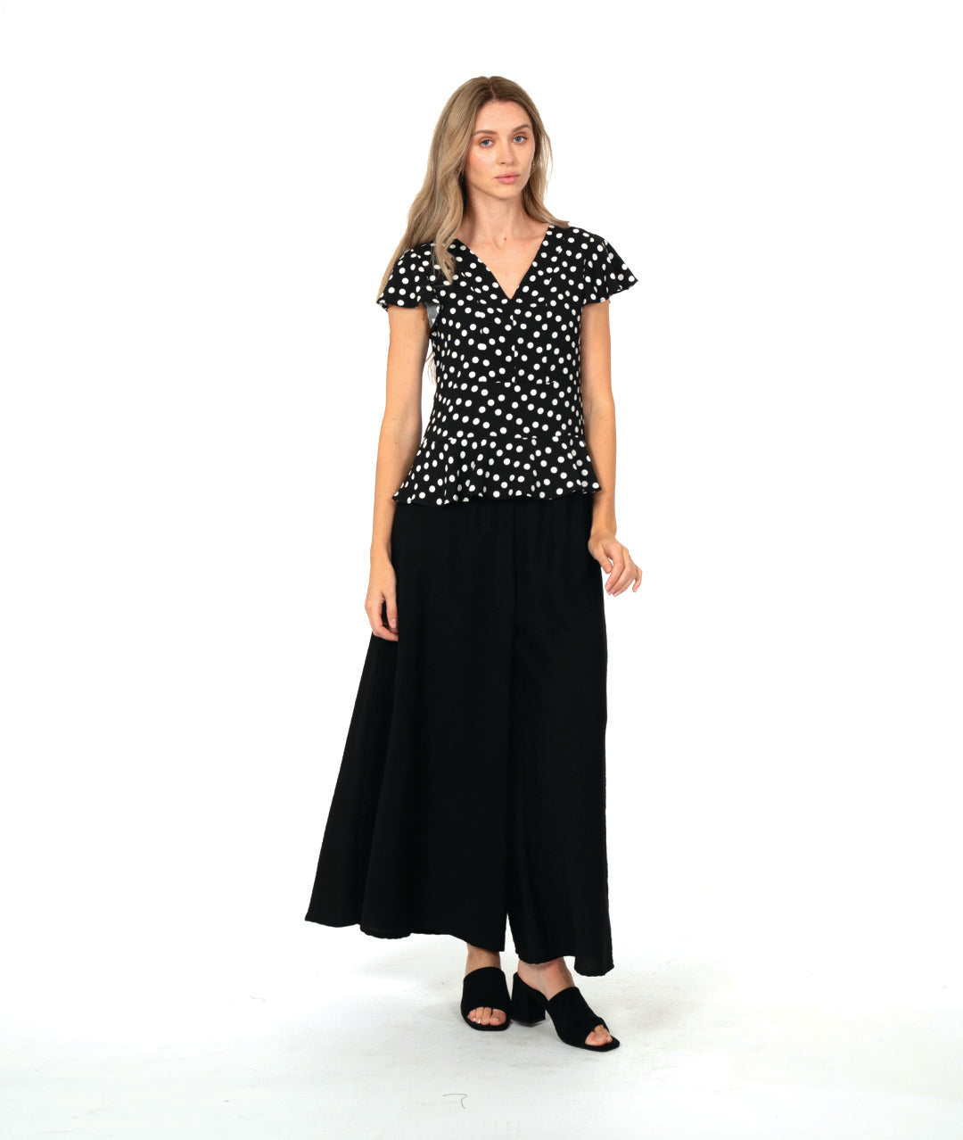 model in a black top with white dots, worn with an ultra wide leg black pant. top is fitted with a v-neck, flutter sleeves and a peplum at the waist