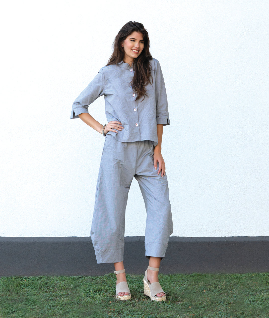 model in a grey and white pinstriped button down blouse with a double collar, oversized cuffs, and contrasted red stitching at the buttons. worn with a matching wide leg pant