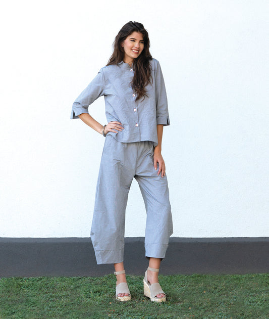 model in a grey and white pinstriped button down blouse with a double collar, oversized cuffs, and contrasted red stitching at the buttons. worn with a matching wide leg pant