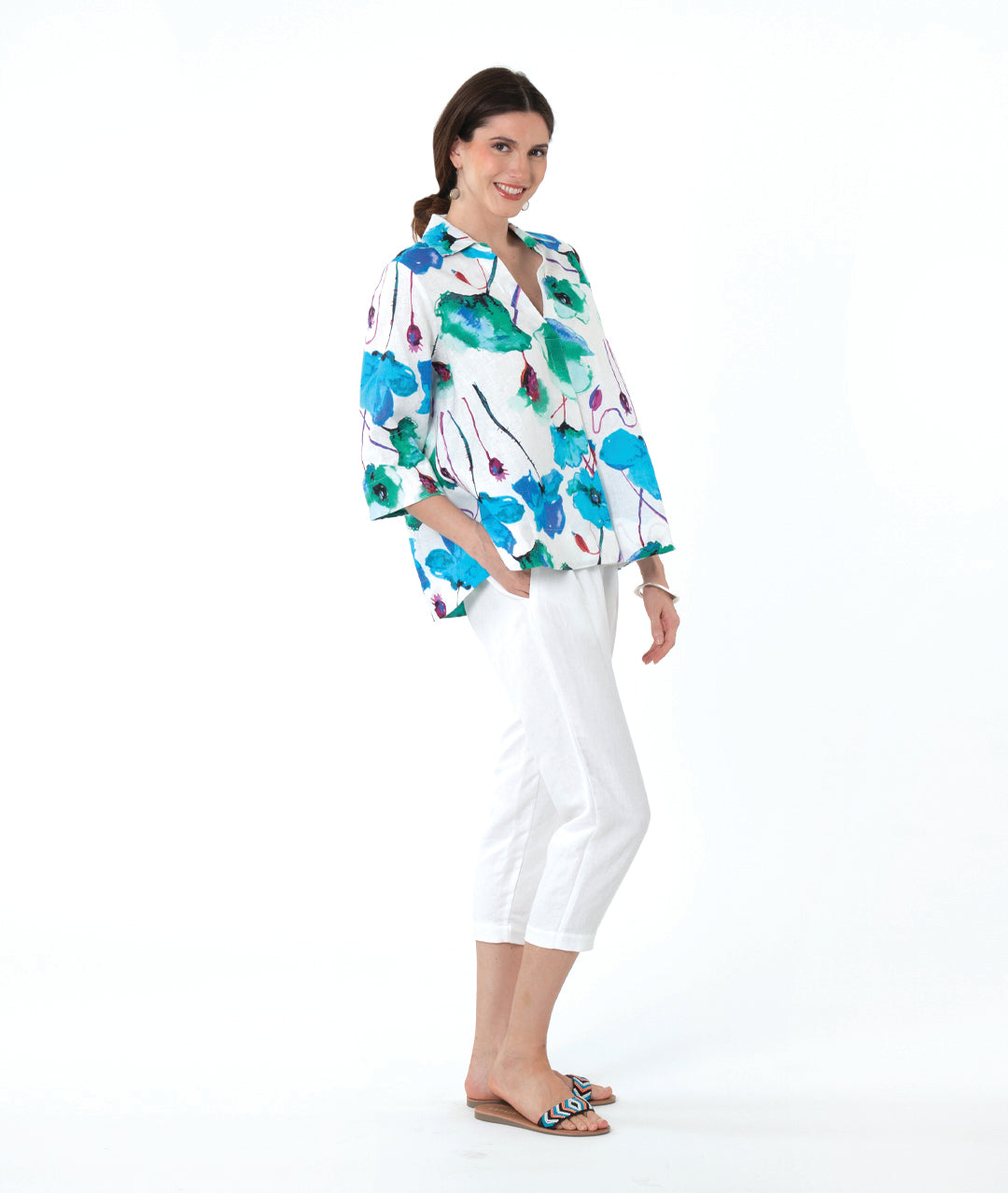 A woman modeling a white three-quarter sleeve button-up shirt with a vibrant green, blue, and purple watercolor floral print, paired with white cropped pants and sandals, against a solid white background.
