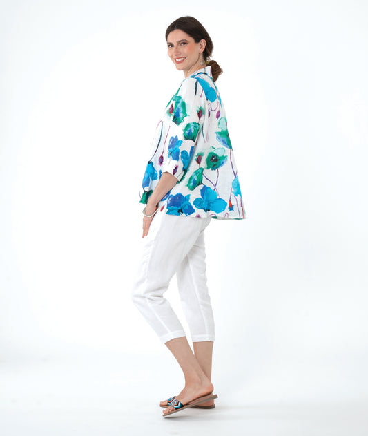 A woman modeling a white three-quarter sleeve button-up shirt with a vibrant green, blue, and purple watercolor floral print, paired with white cropped pants and sandals, against a solid white background.