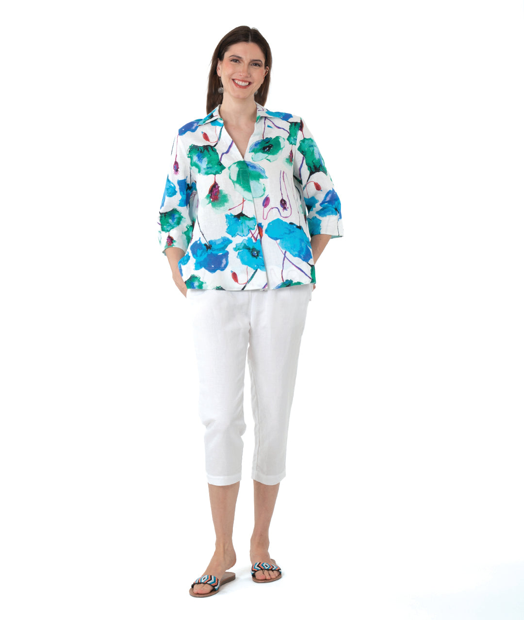 A woman modeling a white three-quarter sleeve button-up shirt with a vibrant green, blue, and purple watercolor floral print, paired with white cropped pants and sandals, against a solid white background.