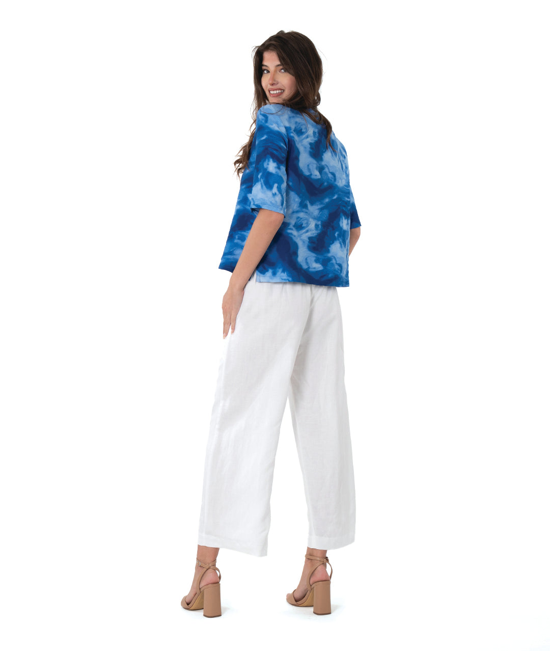 A woman modeling a blue and white abstract patterned short-sleeve top paired with white wide-leg cropped pants and tan heeled sandals, photographed against a seamless white studio background.