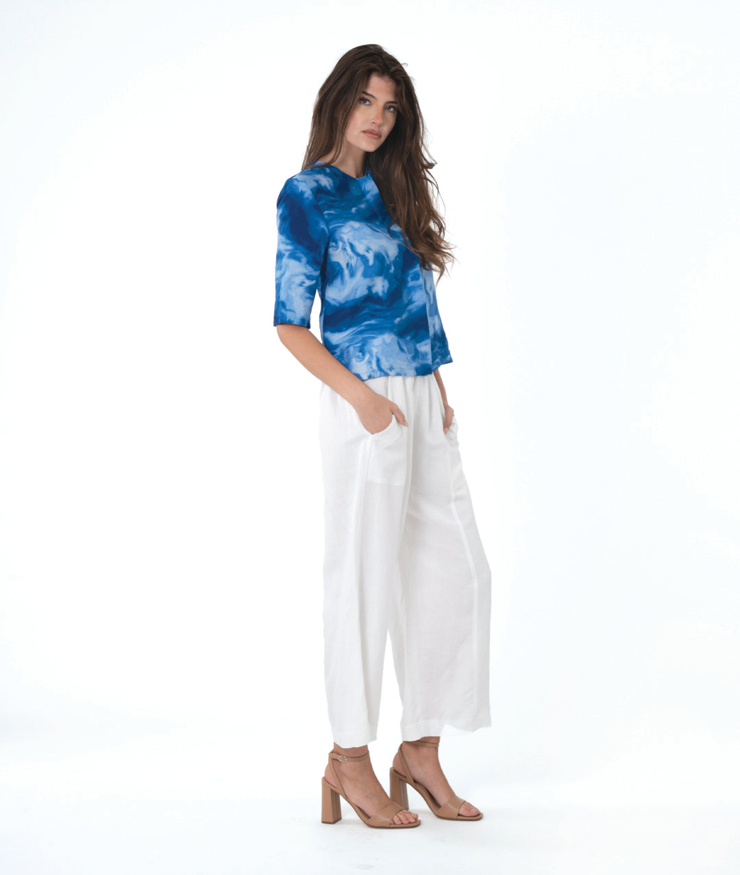 A woman modeling a blue and white abstract patterned short-sleeve top paired with white wide-leg cropped pants and tan heeled sandals, photographed against a seamless white studio background.