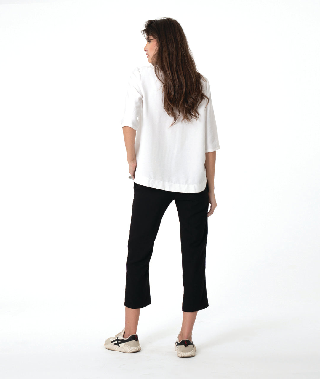 A woman modeling a white V-neck blouse with three-quarter sleeves and black cropped pants, paired with beige sneakers, against a solid white studio background.