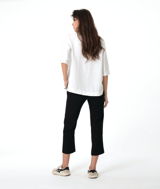 A woman modeling a white V-neck blouse with three-quarter sleeves and black cropped pants, paired with beige sneakers, against a solid white studio background.