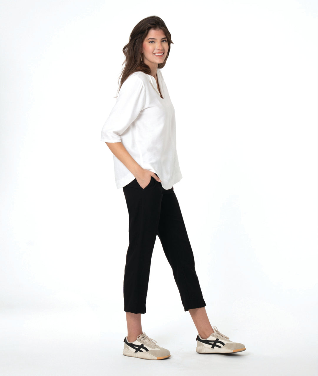 A woman modeling a white V-neck blouse with three-quarter sleeves and black cropped pants, paired with beige sneakers, against a solid white studio background.