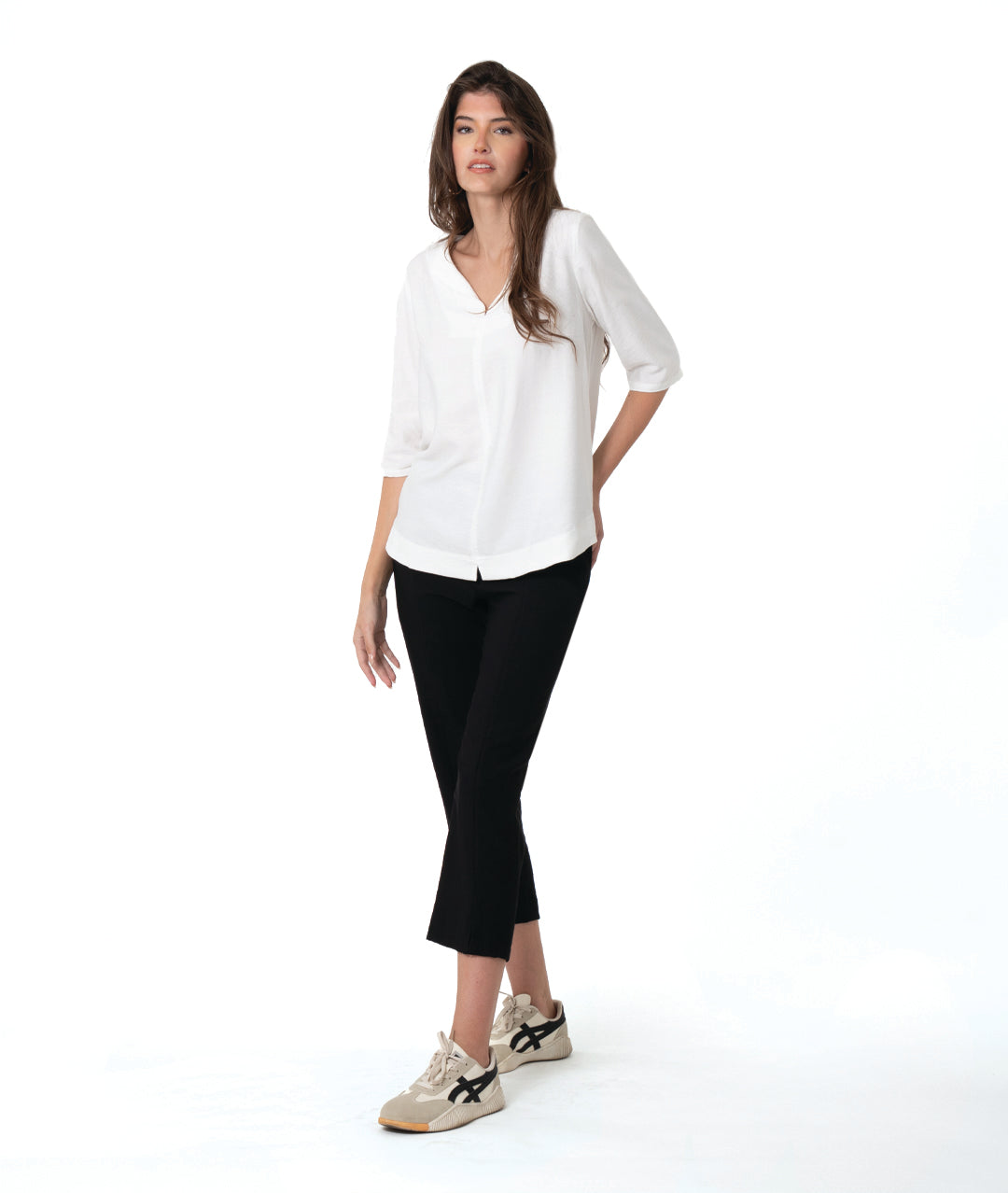 A woman modeling a white V-neck blouse with three-quarter sleeves and black cropped pants, paired with beige sneakers, against a solid white studio background.