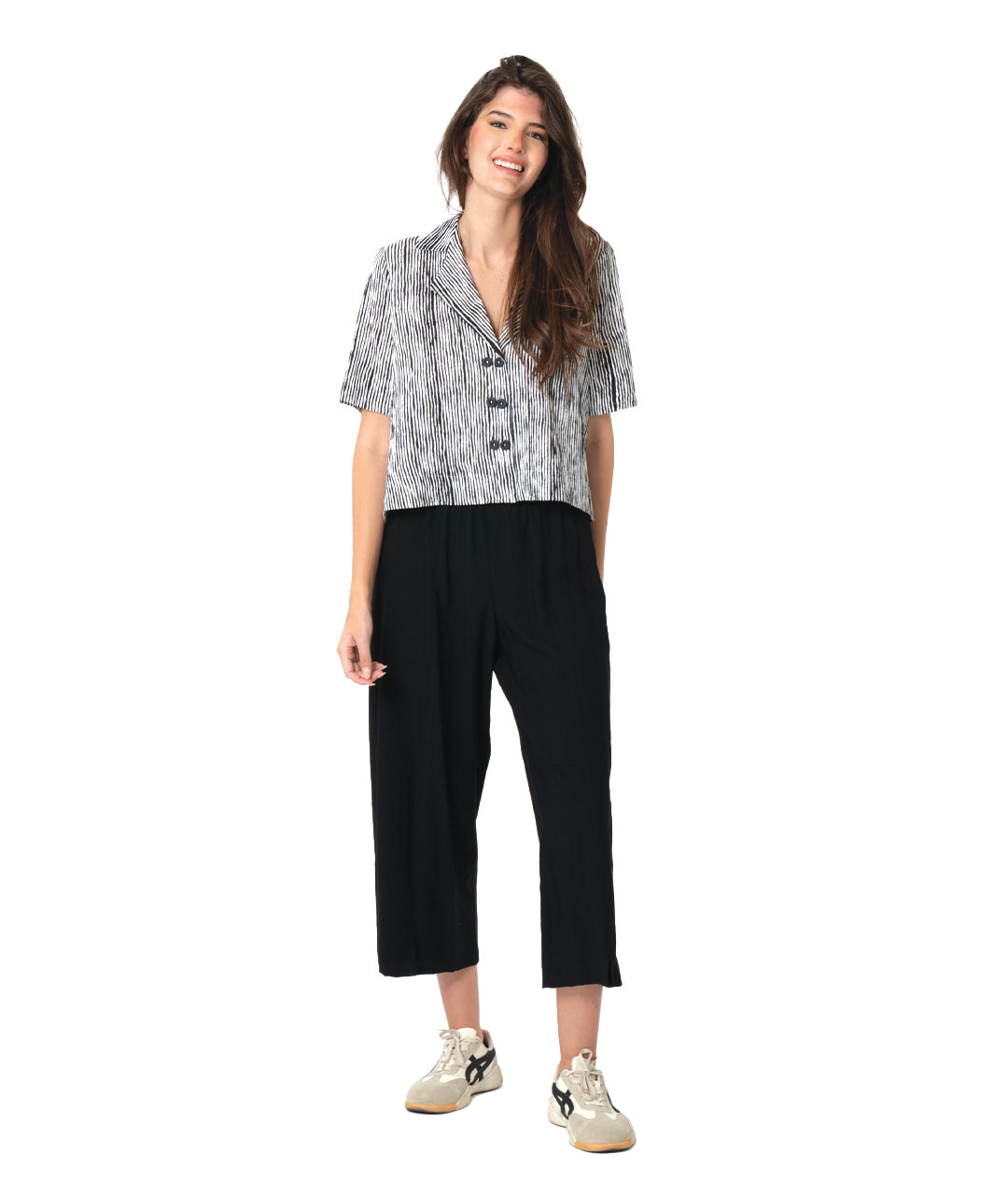 A woman modeling a short-sleeve button-front shirt with a black and white abstract pattern, paired with black cropped pants and beige sneakers, against a plain white background.