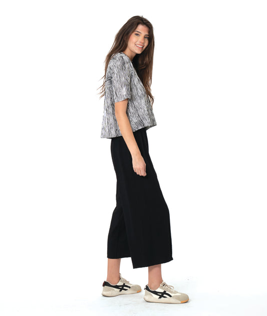 A woman modeling a short-sleeve button-front shirt with a black and white abstract pattern, paired with black cropped pants and beige sneakers, against a plain white background.