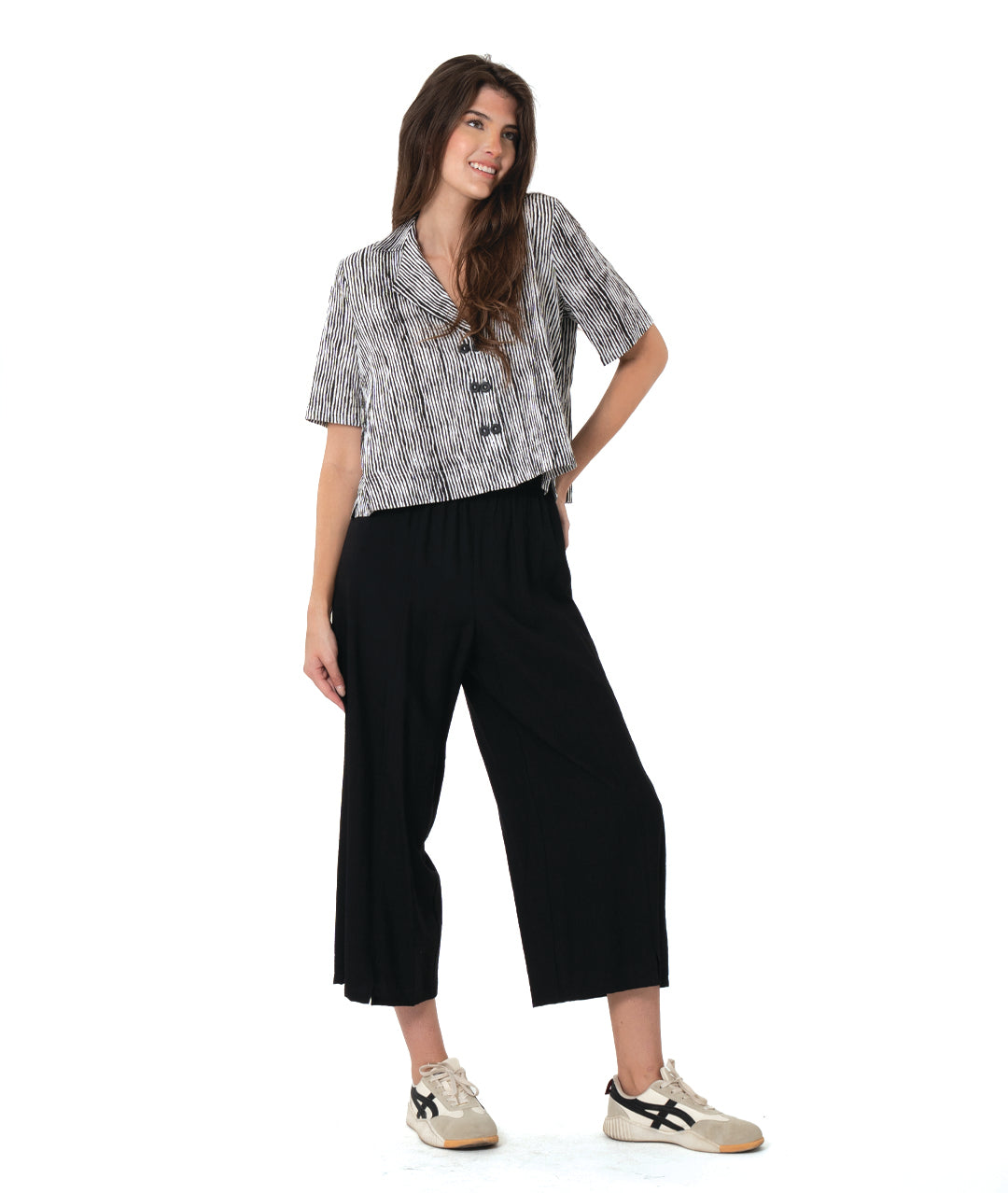 A woman modeling a short-sleeve button-front shirt with a black and white abstract pattern, paired with black cropped pants and beige sneakers, against a plain white background.