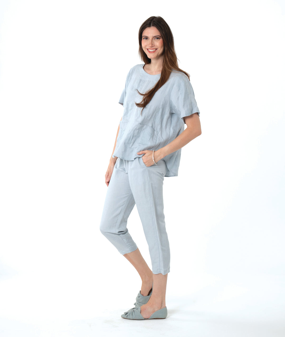 A woman modeling a light blue linen two-piece lounge set consisting of a short-sleeve top and cropped pants, paired with matching flat shoes, against a solid white background.