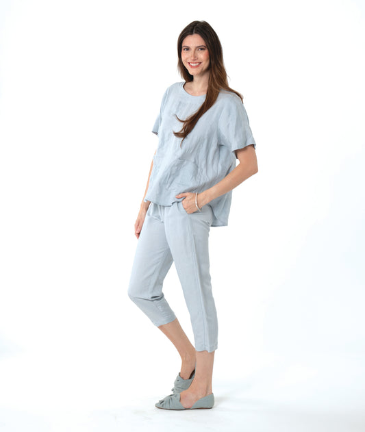 A woman modeling a light blue linen two-piece lounge set consisting of a short-sleeve top and cropped pants, paired with matching flat shoes, against a solid white background.