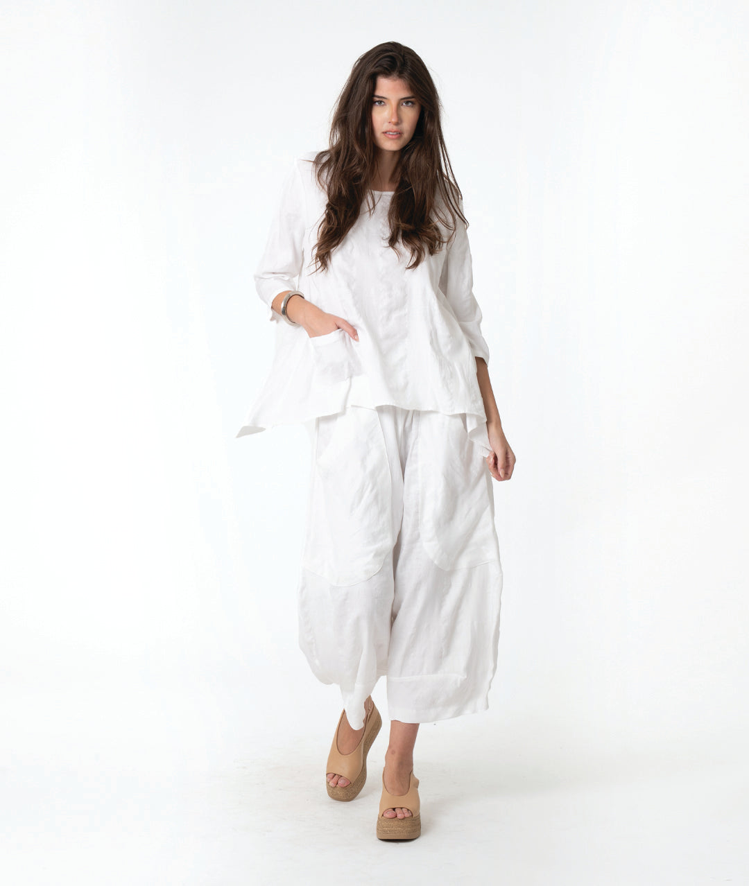 A woman modeling a white two-piece linen outfit consisting of a loose long-sleeve top and matching wide-leg cropped pants, paired with tan wedge sandals, against a plain white studio background.