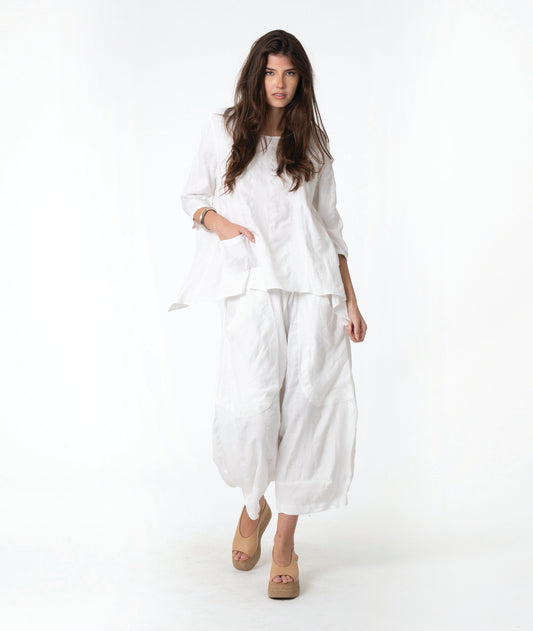 A woman modeling a white two-piece linen outfit consisting of a loose long-sleeve top and matching wide-leg cropped pants, paired with tan wedge sandals, against a plain white studio background.
