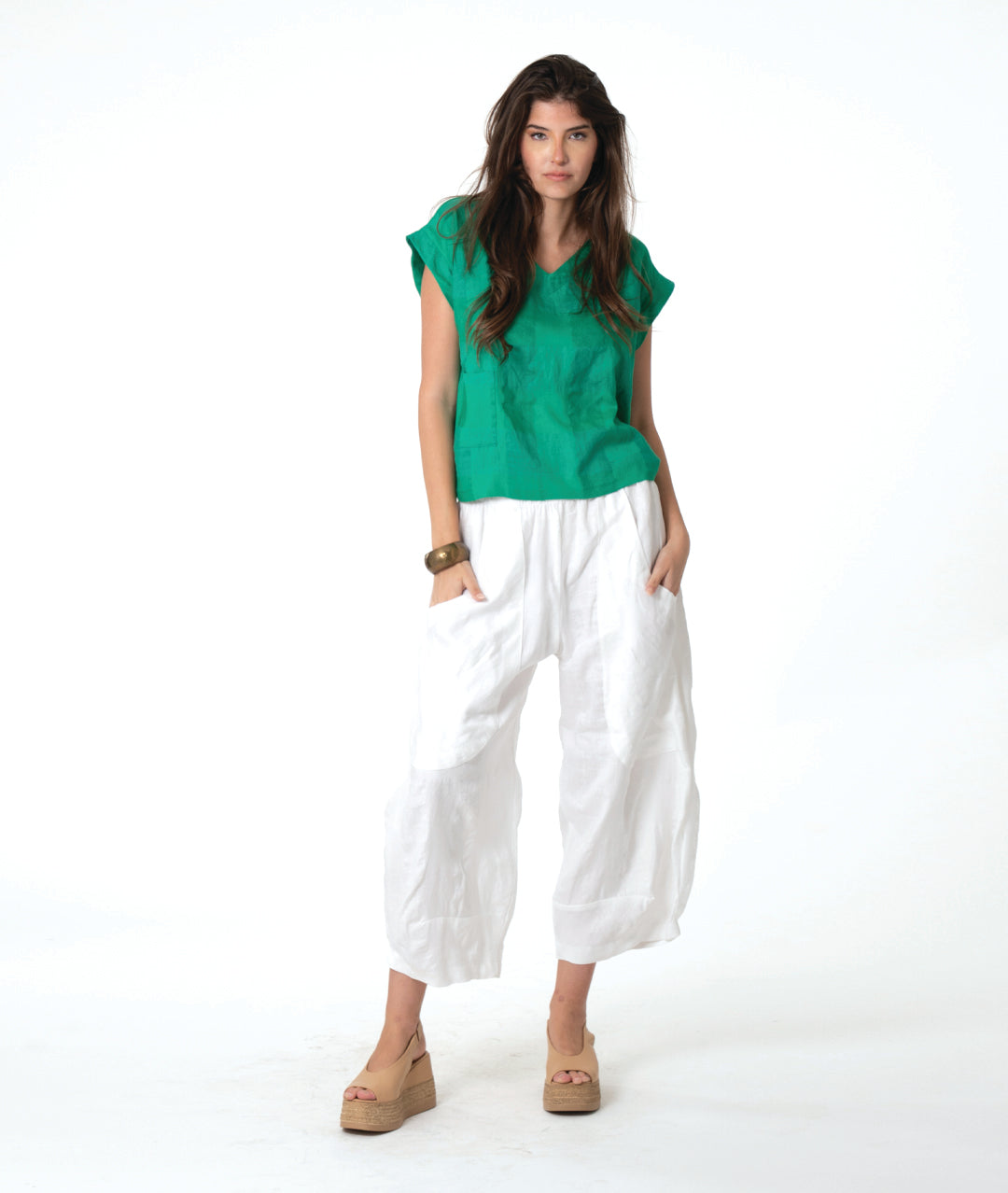 A woman modeling a vibrant green short-sleeve top paired with loose white cropped pants and tan wedge sandals, set against a plain white studio background.