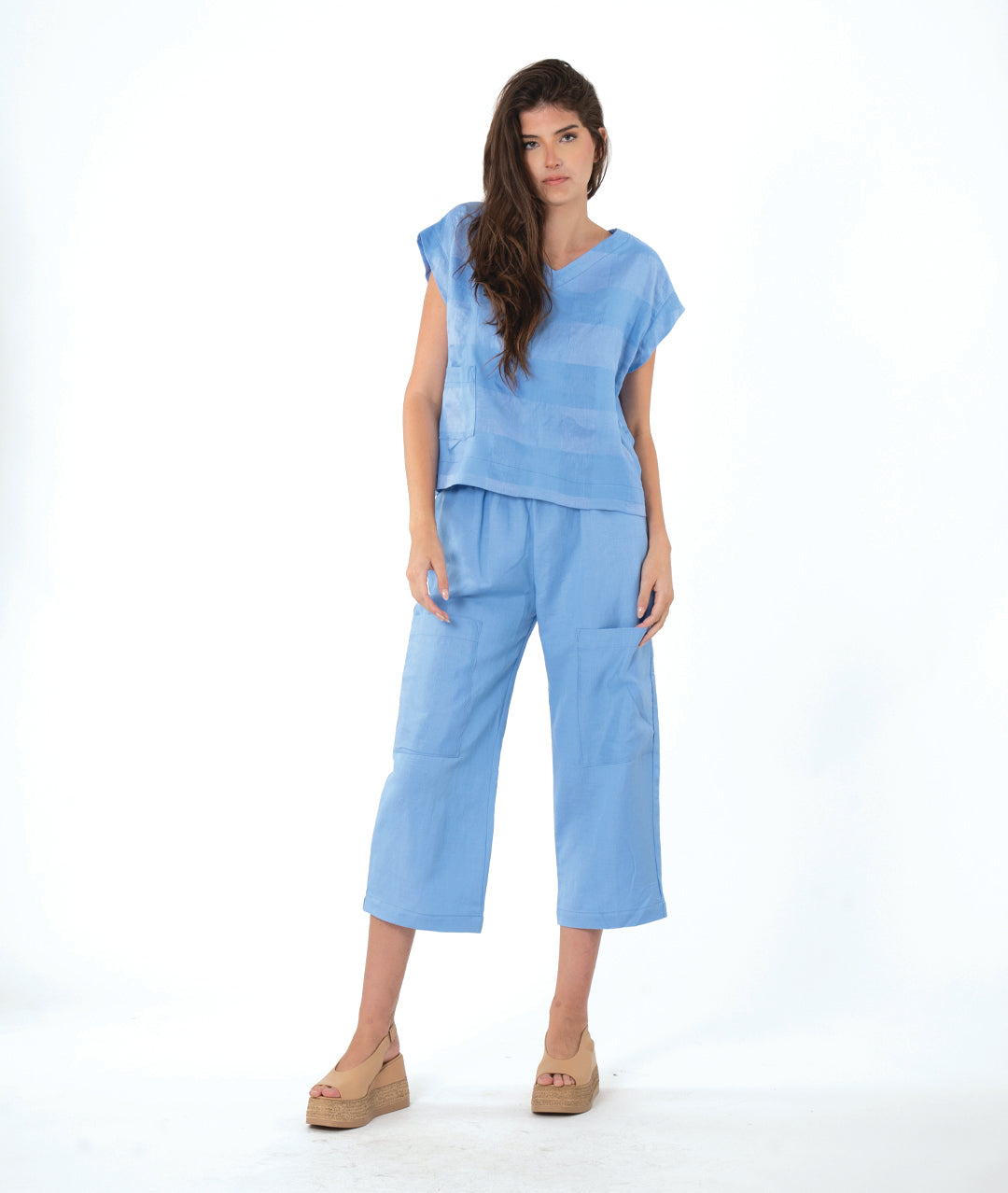 A woman modeling a light blue two-piece linen set consisting of a short-sleeve V-neck top and matching cropped pants, paired with tan wedge sandals, against a white studio background.