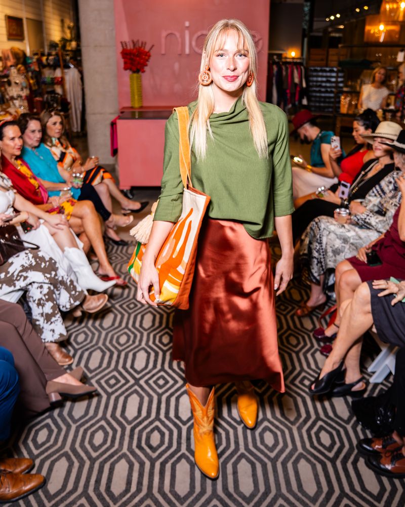 model posing on a runway wearing green cowl top, copper satin skirt, yellow cowboy boots and a desert scene tote bag