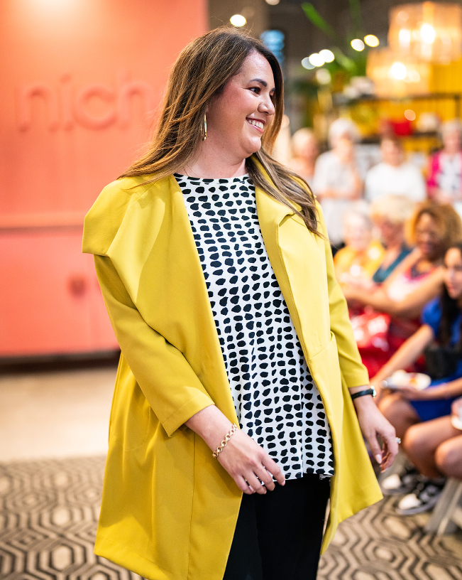 model posing for people wearing a gold asymmetrical jacket with a large collar and black and white polka dot top underneath.