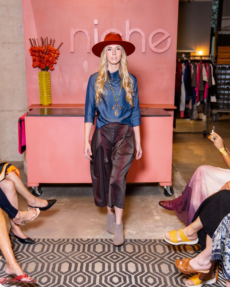 model wearing orange hat, blue cowl top, statement necklace, brown shimmer stripe pants with taupe boots on runway