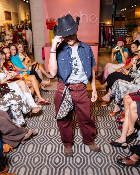model wearing short sleeve navy pearl snap shirt with grey tshirt underneath, burgundy pants, black cowboy hat and brown cowboy boots on a runway