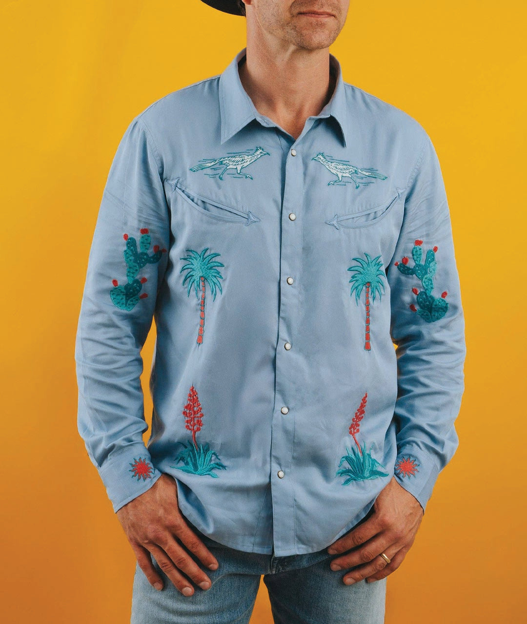 Man wearing a light blue embroidered shirt with palm trees and cacti on an orange background