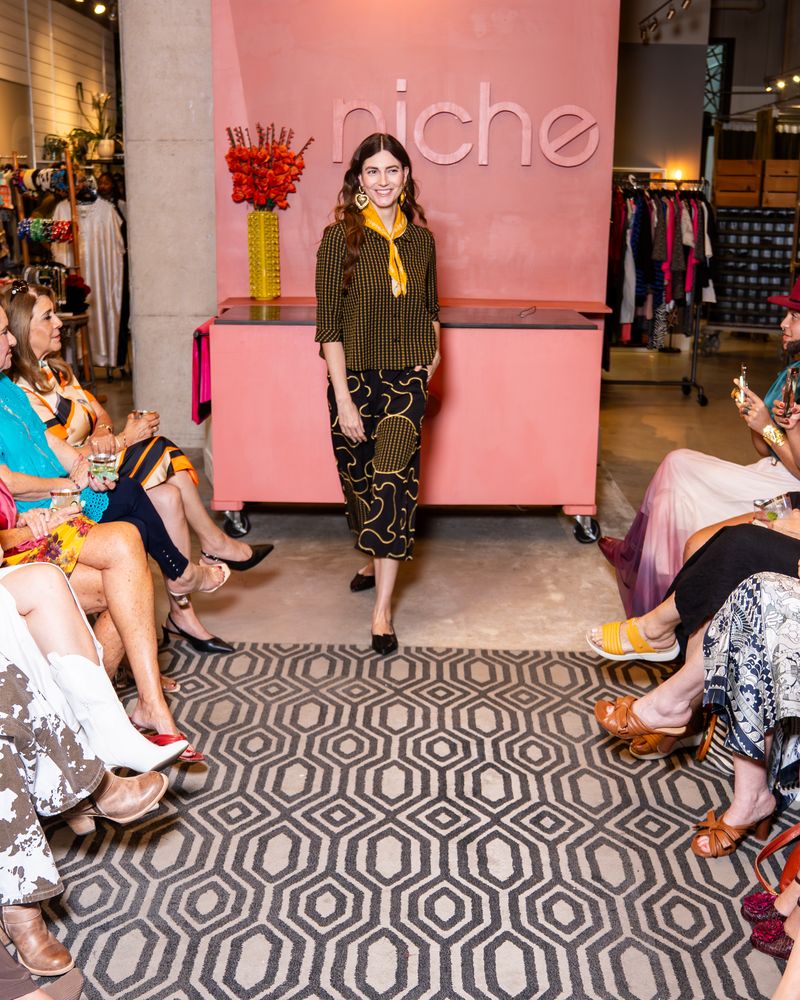 Model walking on a catwalk at a fashion event with wearing yellow check button up top, swirl pattern pants and yellow bandana
