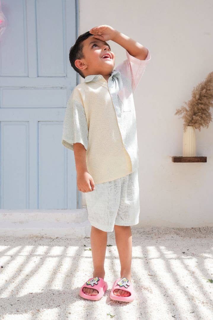 Child wearing a light-colored outfit with a white shirt and shorts, standing on a textured surface with a light blue wall in the background.