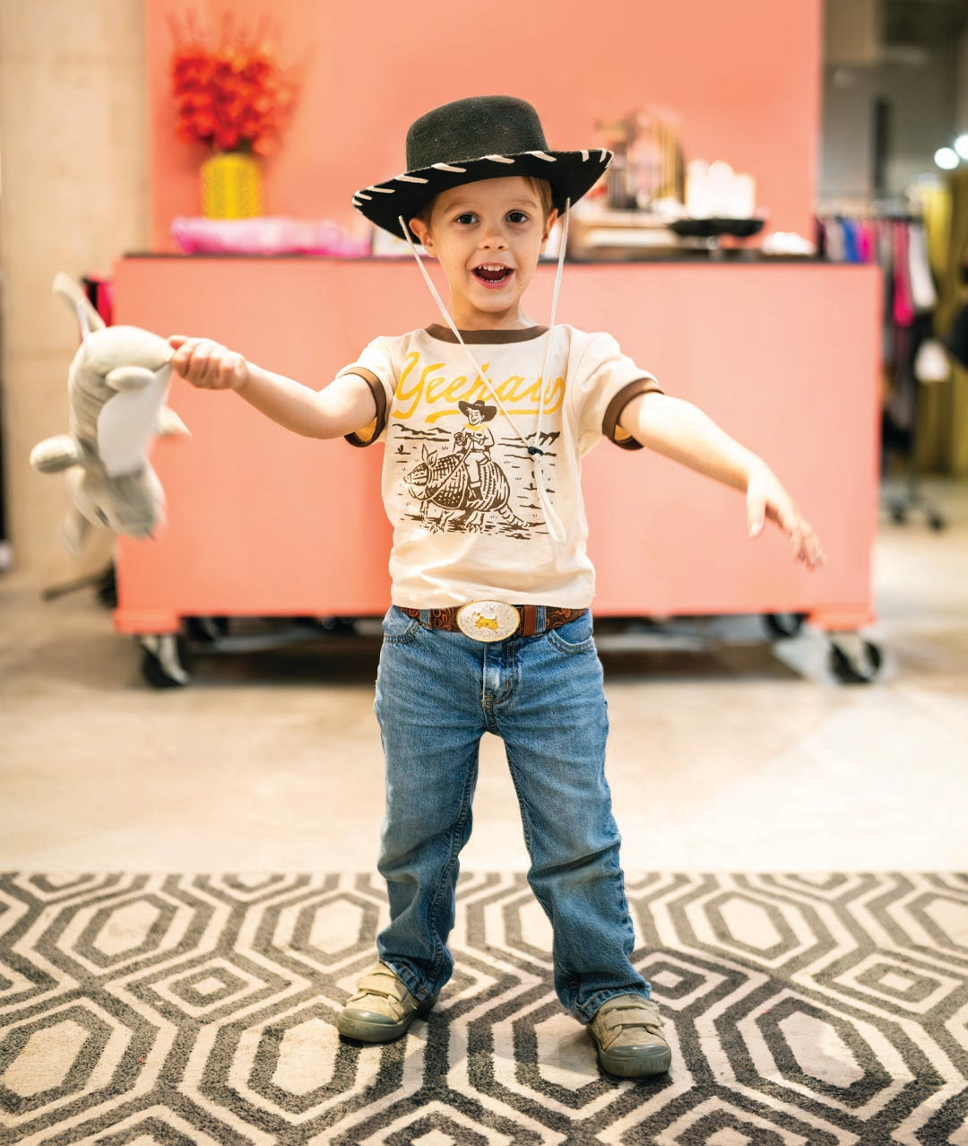 Child in cowboy hat and jeans holding a toy, standing on a patterned rug with a pink wall in the background.