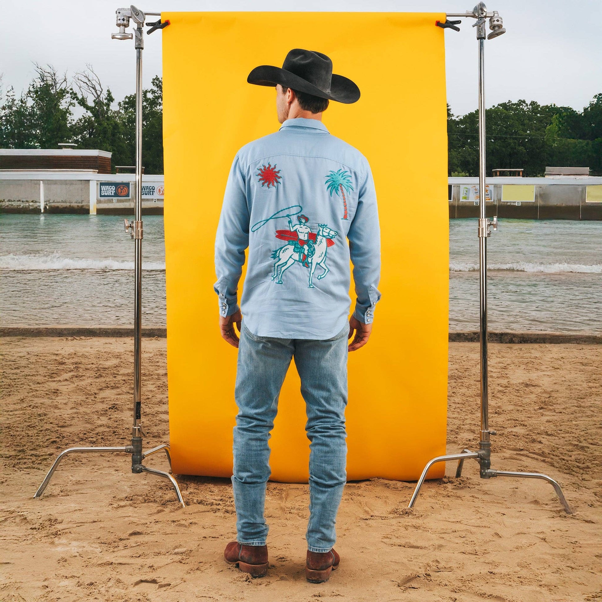 Person wearing a cowboy hat and denim outfit with a colorful design on a yellow backdrop in an outdoor setting.