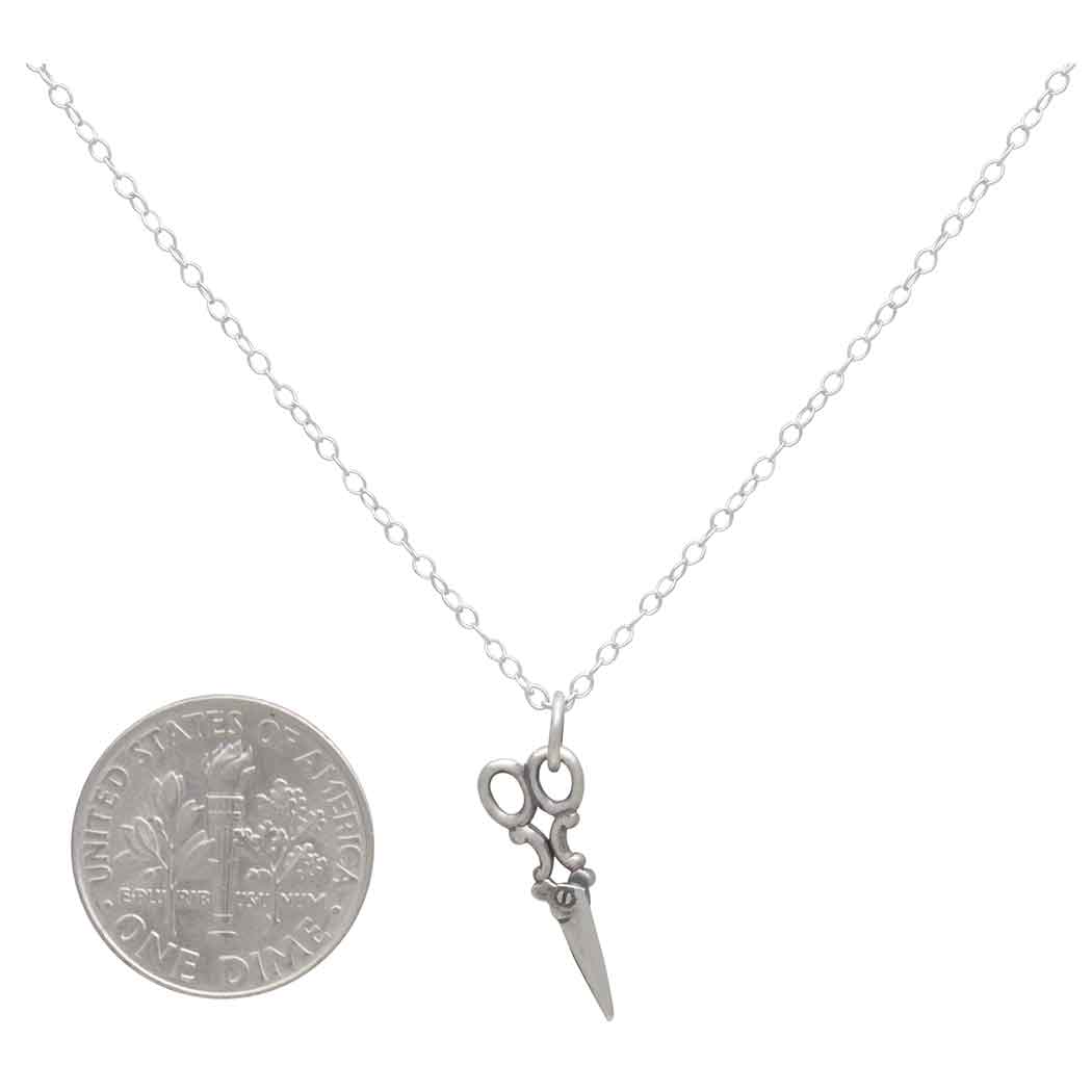 Silver necklace with a pair of scissors and dagger pendant next to a U.S. dime coin on a white background