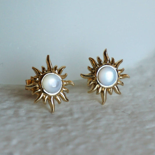 Gold sun-shaped earrings with pearl center on a light background.
