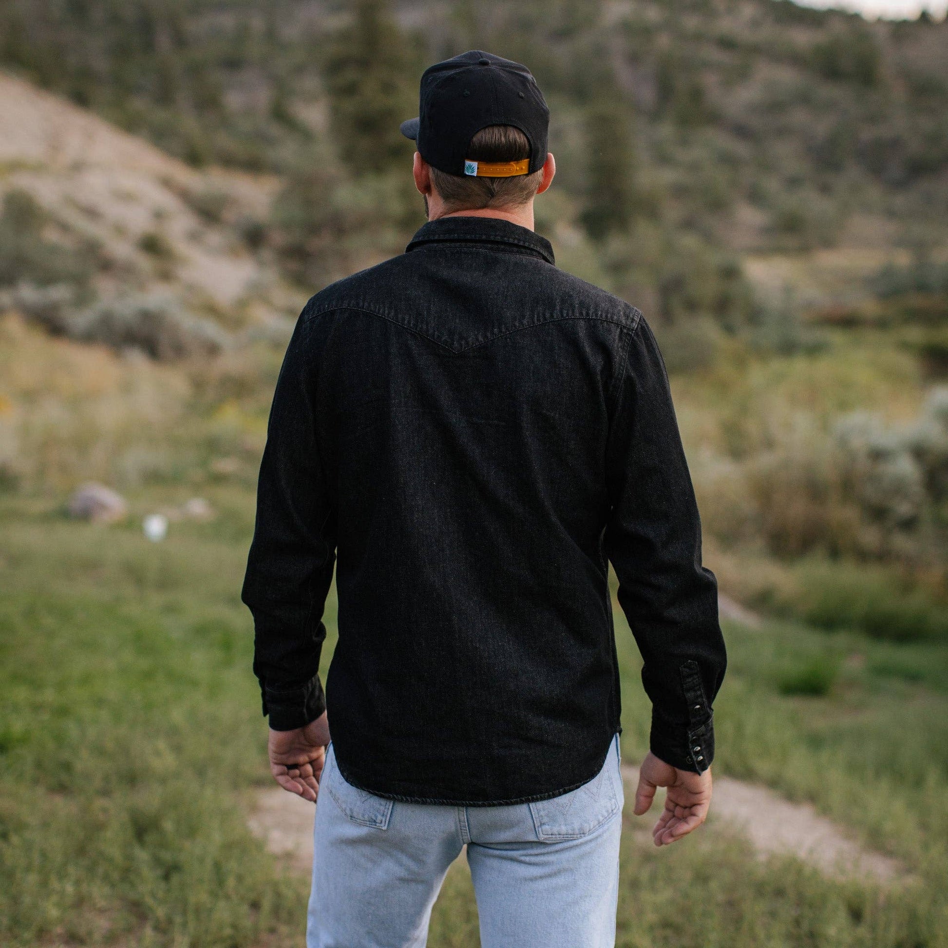 back side of male model wearing long sleeve black denim shirt, jeans and hat standing outside in nature