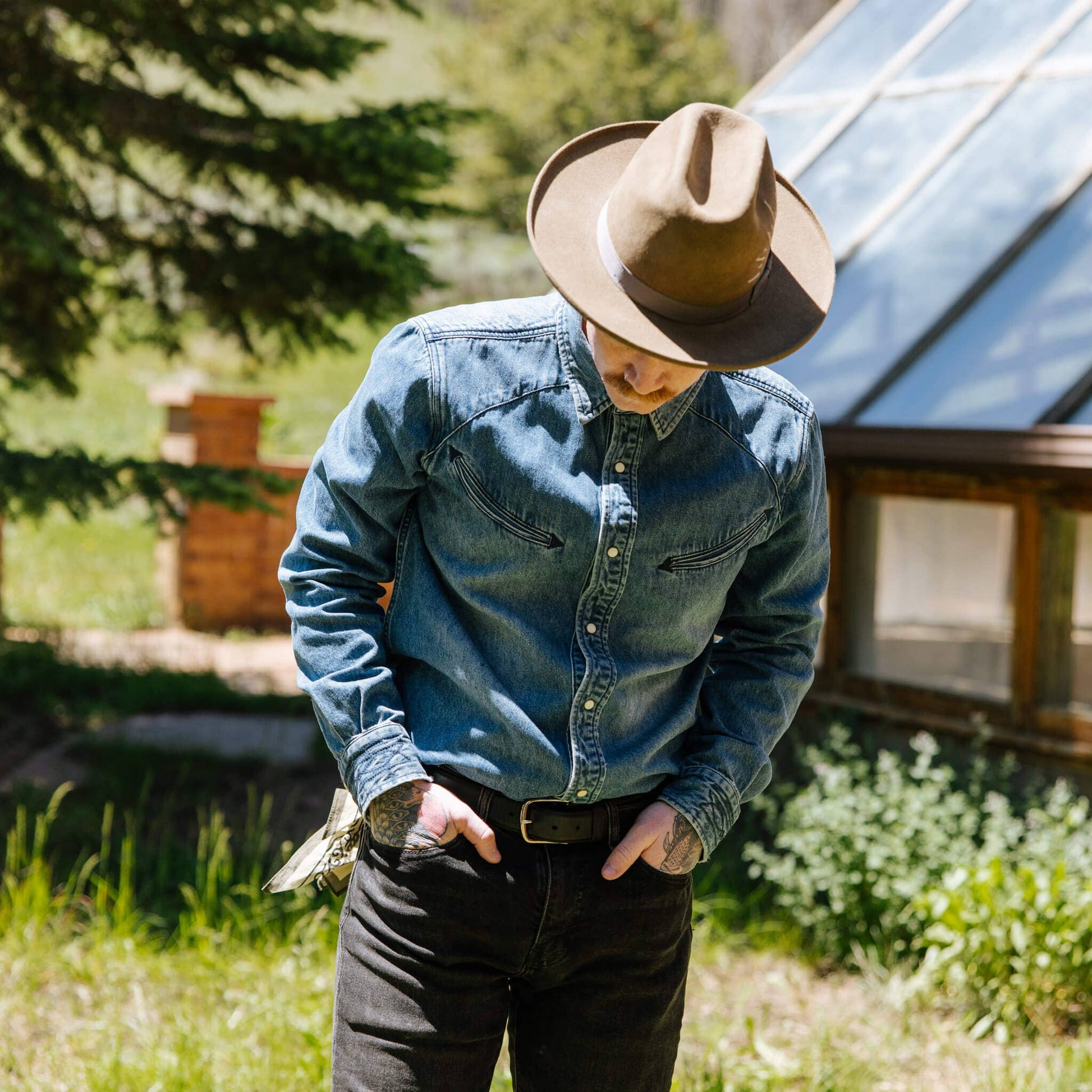 model wearing indigo denim collared long sleeve, with arrow stitching pockets and pearl snaps. worn with brown hat and black pants.