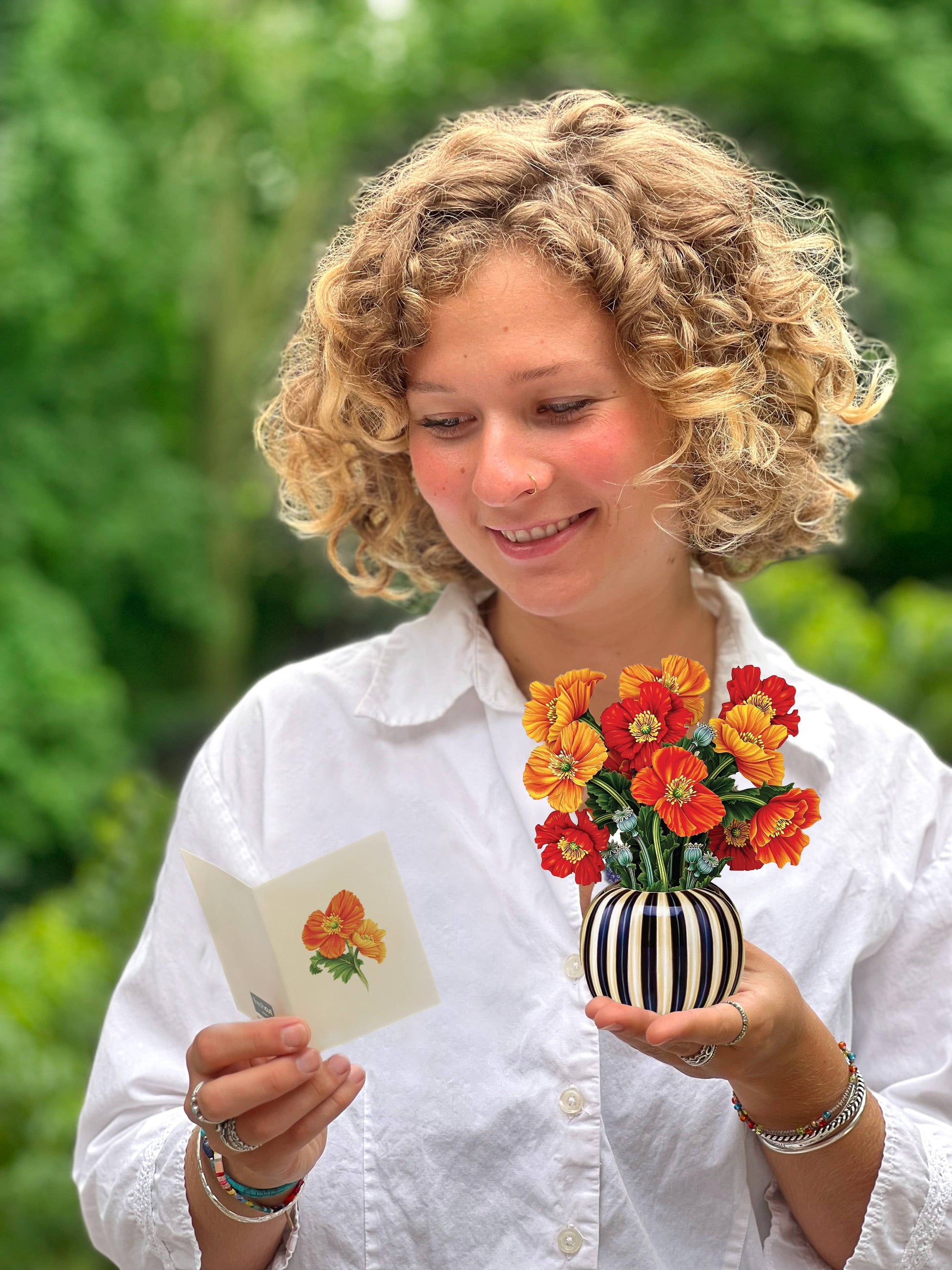 Person holding a small vase with flowers and a matching card outdoors.