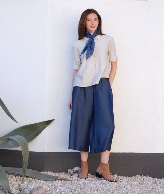 Woman wearing a white blouse with a blue scarf and blue pants standing against a white wall with a plant in the foreground.