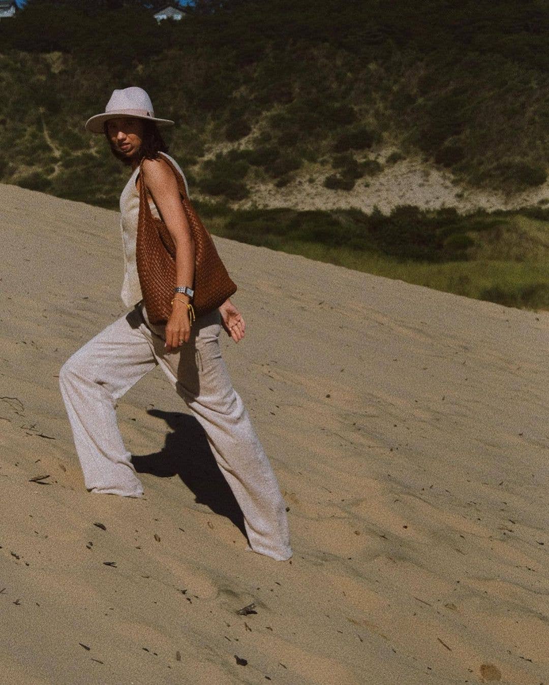 Person walking on a sandy dune wearing a hat, beige outfit, and brown bag.