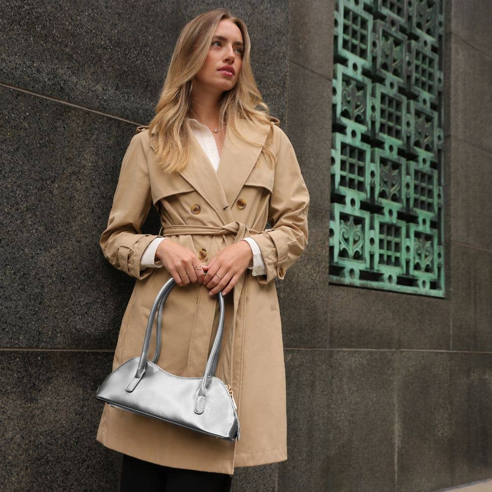Woman in a beige trench coat holding a silver handbag against a textured wall.