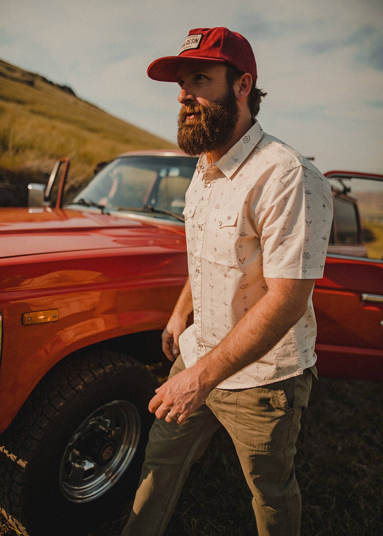 model wearing light beige western short sleeve shirt with small patterns, double chest pocket and top entry button closure, and pearl snap buttons, with red hat and truck in background