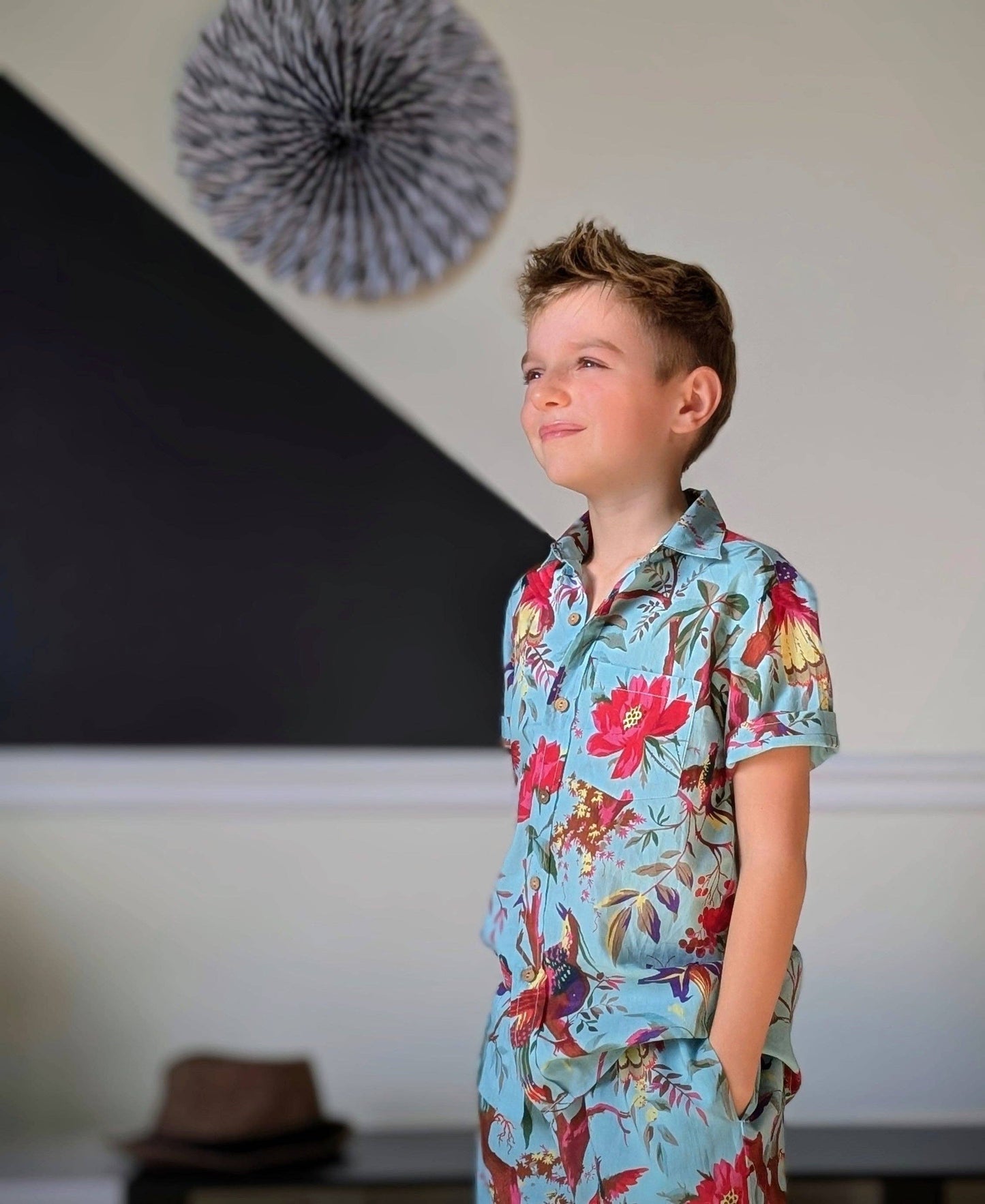 Child wearing a colorful floral shirt standing in a room with a decorative wall hanging.