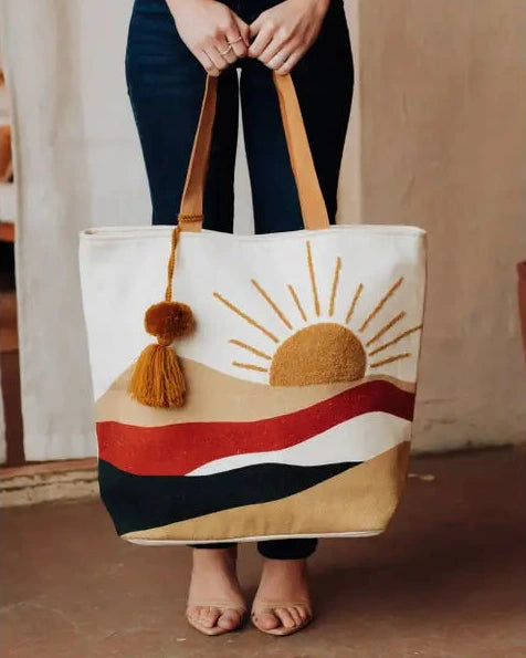 Person holding a tote bag with a sunset design and tassels.