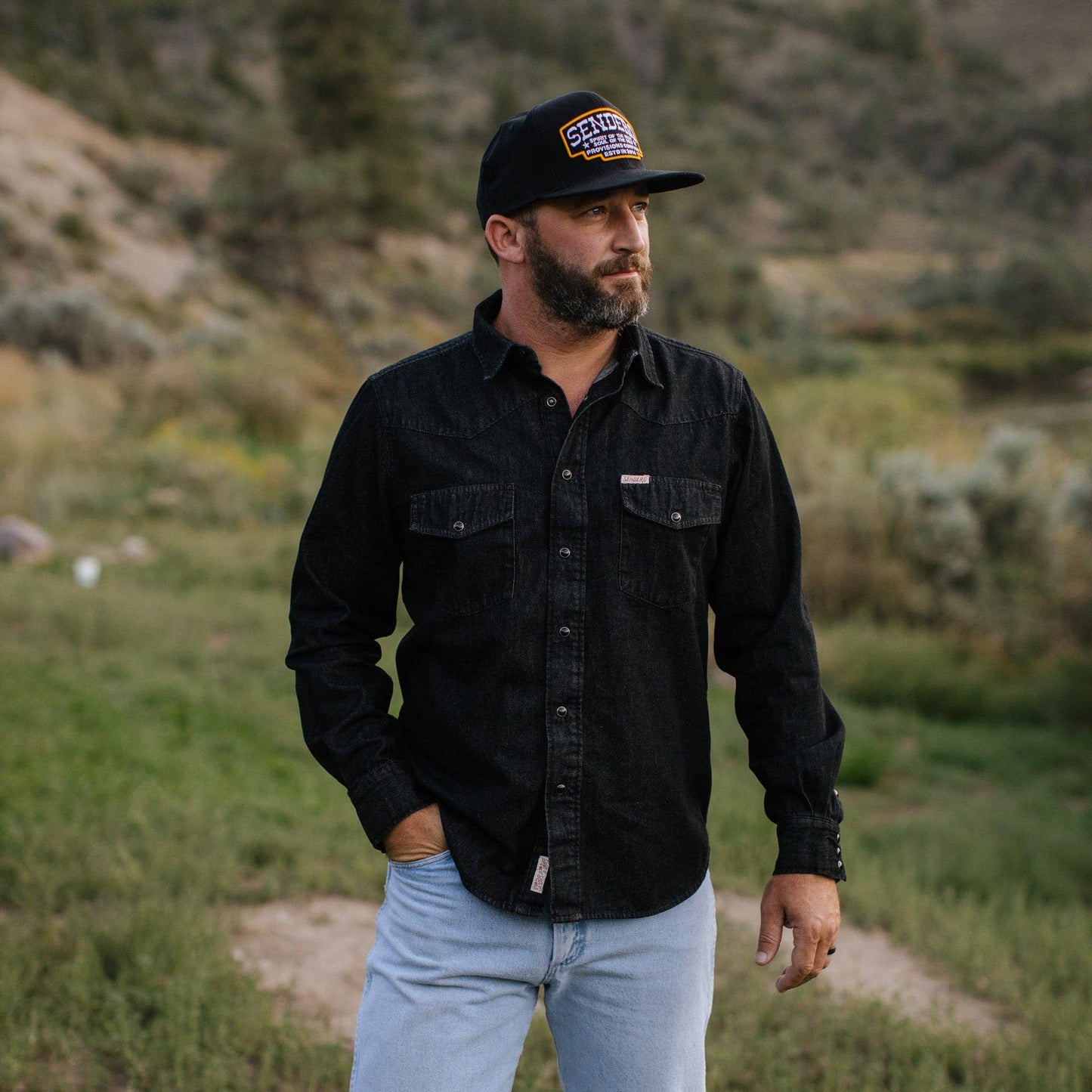 male model wearing long sleeve black denim shirt, jeans and hat standing outside in nature