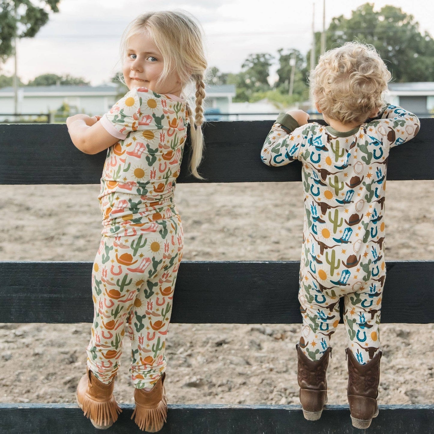 Two children wearing patterned rompers standing on a fence with a rural background.
