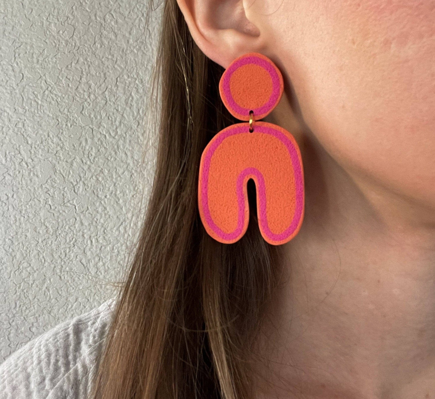 Clay post earrings in pink and orange in circle and u-shape. on a close up on a brown haired model and a white wall