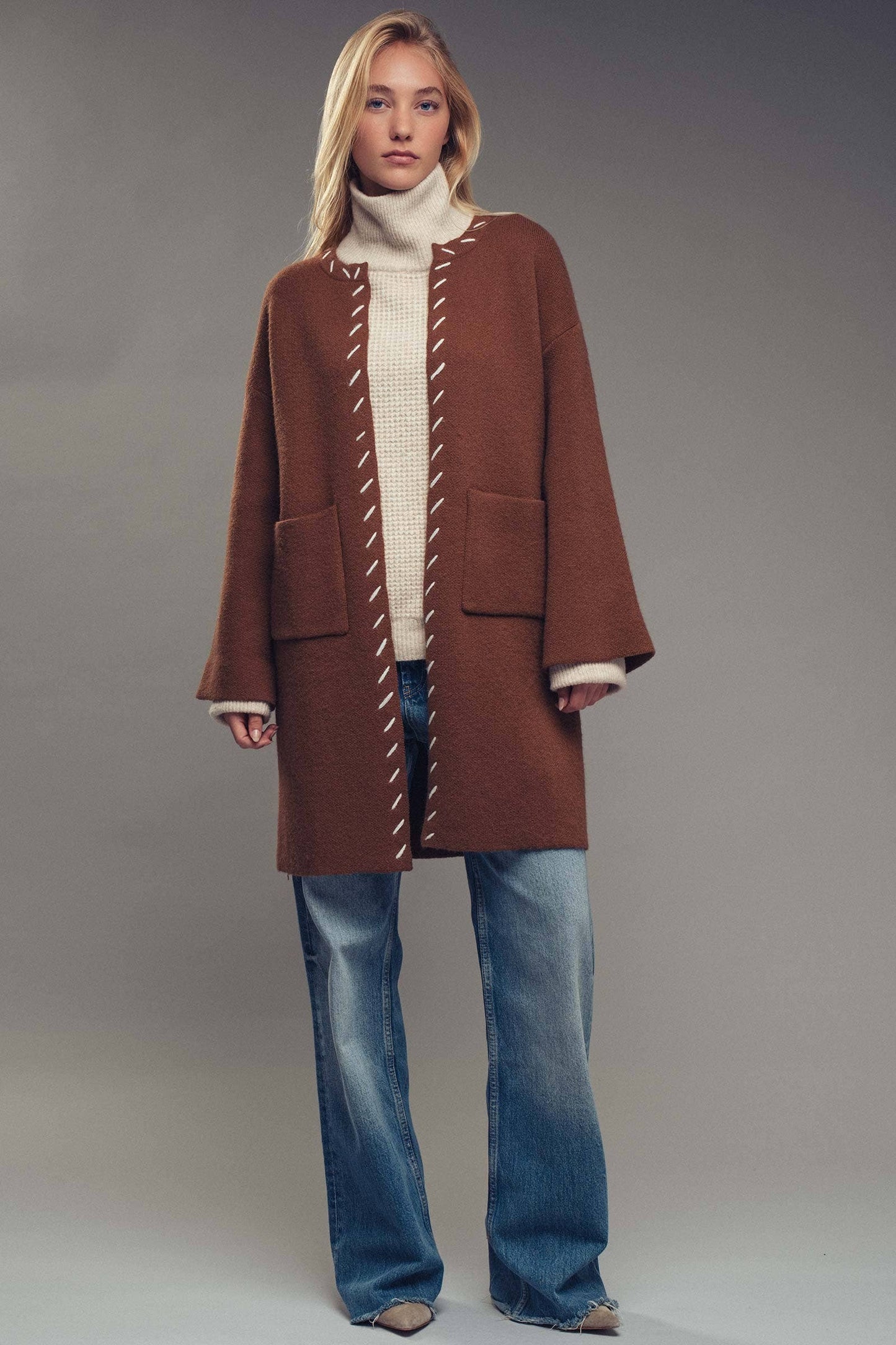 Woman wearing a brown coat with white stitching, beige turtleneck, and blue jeans on a gray background